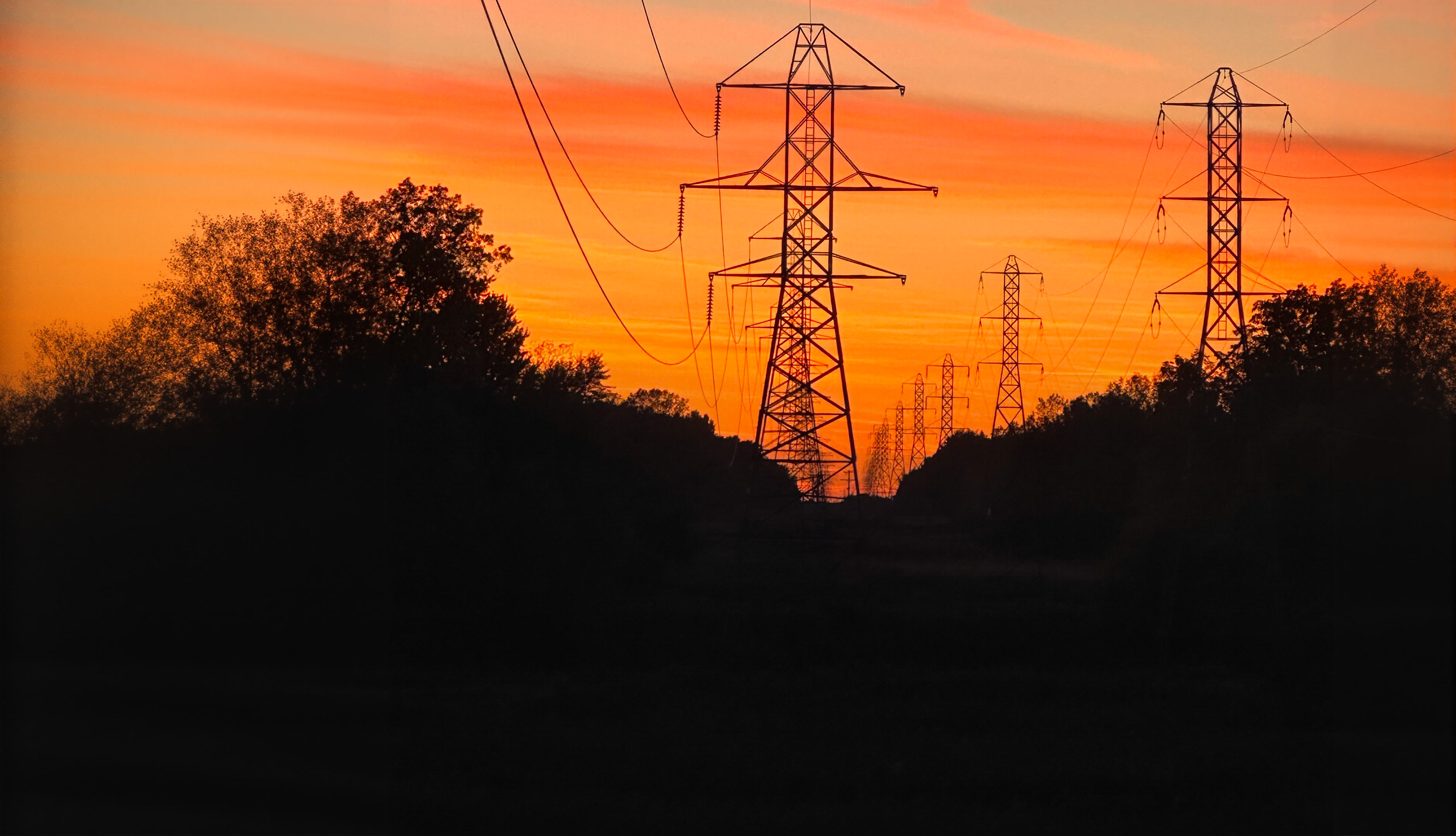 Steel power transmission towers silhouetted against a brilliant orange sunset sky.